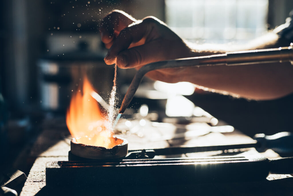 Closeup of a jeweler using a torch to melt metal in a crucible while working in his jewelry design studio